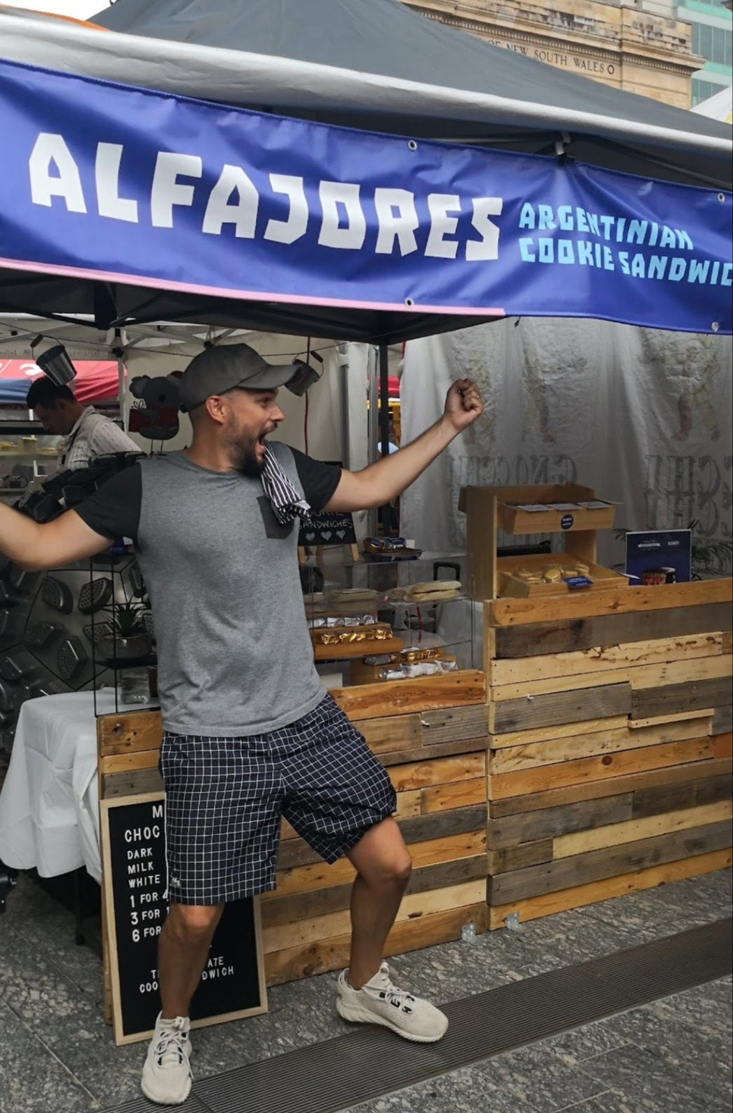 Man standing under a blue banner with 'Alfajores' text at an outdoor market stall.